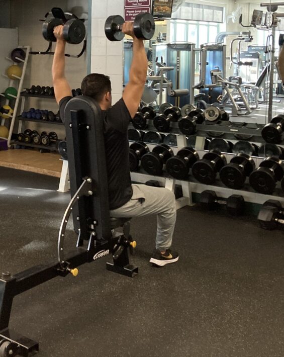 Picture of man from behind sitting on a weight bench lifting two dumbbells above his head
